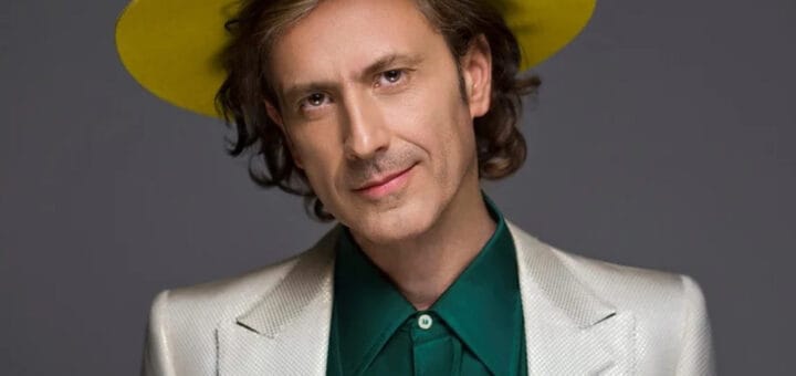 alt="Coti con saco claro camisa verde y sombrero amarillo"