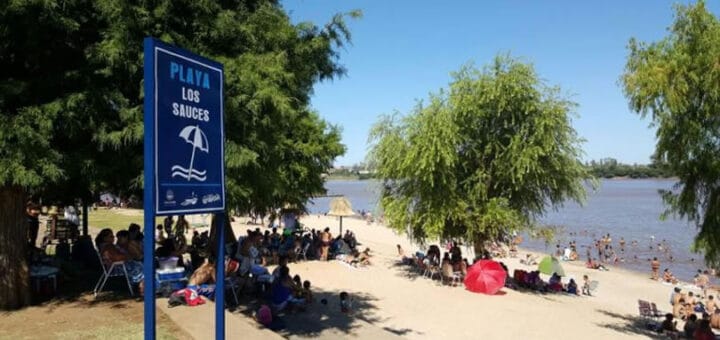 alt="playa los sauces con gente en la arena y bajo los árboles y cartel indicador"