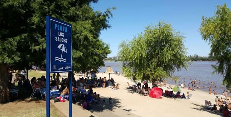 alt="playa los sauces con gente en la arena y bajo los árboles y cartel indicador"