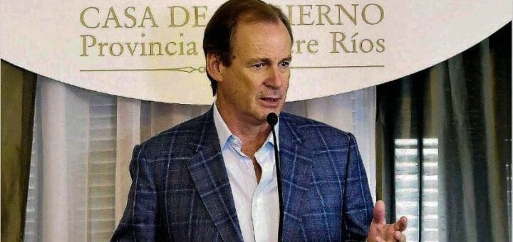 alt="el gobernador Gustavo Bordet de saco a cuadros y camisa blanca habla frente a un micrófono, detrás aparece un cartel que dice casa de gobierno provincia de entre rios"