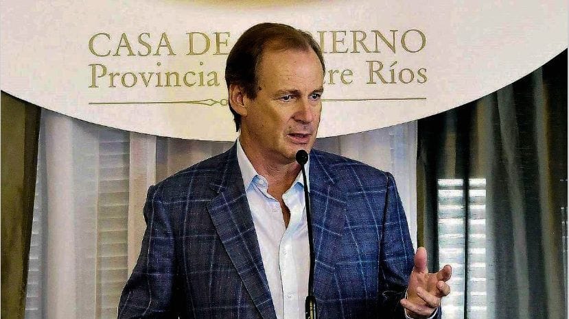 alt="el gobernador Gustavo Bordet de saco a cuadros y camisa blanca habla frente a un micrófono, detrás aparece un cartel que dice casa de gobierno provincia de entre rios"