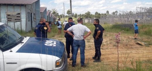 alt="tres policías, un funcionario municipal, un patrullero, casas y, más atrás, gente que participó en el intento de usurpación de viviendas"