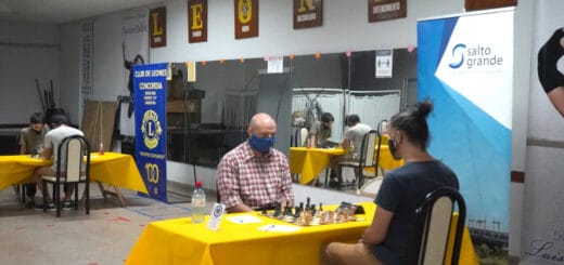 alt="en primer plano, dos ajedrecistas jugando al lado de un cartel de Salto Grande, en segundo plano otros dos ajedrecistas junto a un cartel del club de leones"