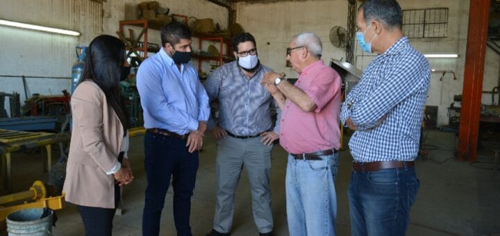 alt="una mujer y cuatro hombres en semicírculo, dialogan en el interior de un galpón"