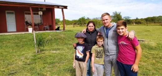 alt="en la imagen, una familia compuesta por un matrimonio y tres niños; están en el campo, detrás de ellos una vivienda de madera"