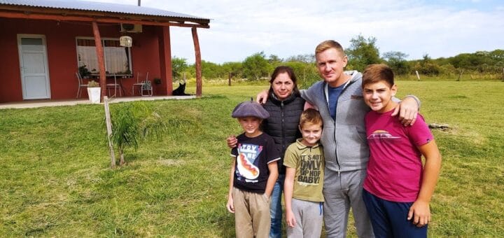 alt="en la imagen, una familia compuesta por un matrimonio y tres niños; están en el campo, detrás de ellos una vivienda de madera"
