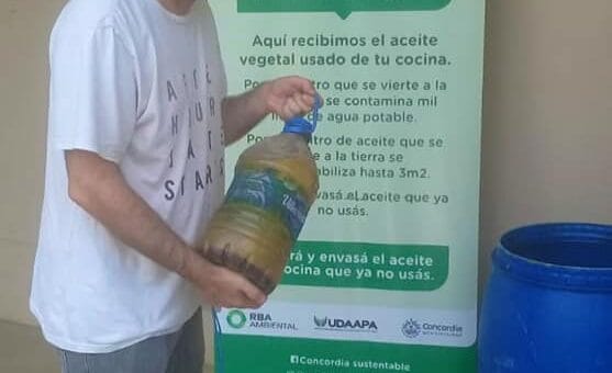alt="en la imagen, una persona sostiene una botella con aceite vegetal usado, también hay un tonel y un bidón en el piso, detrás de todo un cartel"