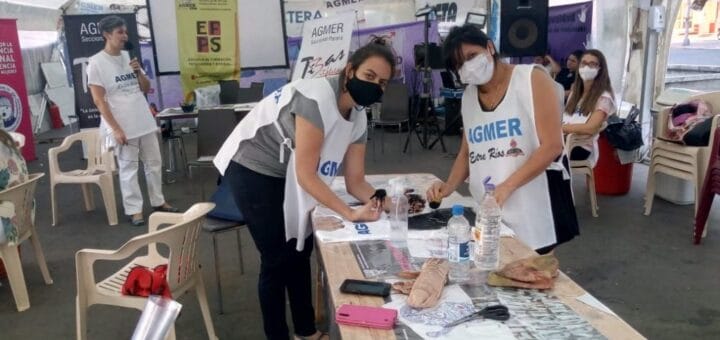 alt="dos mujeres con ropa con inscripciones de AGMER preparan elementos diversos sobre una mesa, más atrás hay otras dos mujeres"