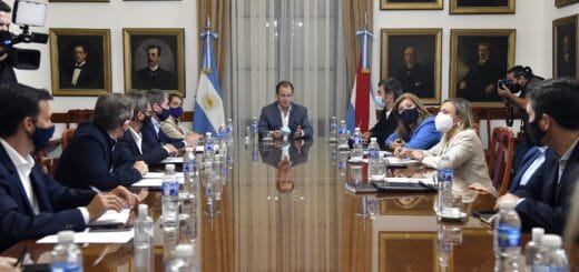 alt="en la imagen, una mesa larga, en una cabecera el gobernador, a sus lados una bandera argentina y otra de entre rios; a ambos lados de la mesa, varios funcionarios sentados; detrás de todos, una ventana con largas cortinas y dos paredes llenas de retratos"