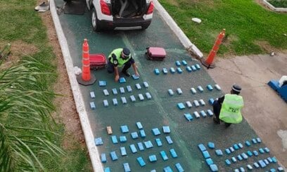 alt="en la parte inferior de la fotografía, dos policías ordenan decenas de teléfonos celulares sobre el pavimiento; en la parte superior hay un automóvil de color blanco, con las puertas y el baúl abiertos, a la derecha una palmera pequeña"