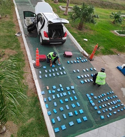 alt="en la parte inferior de la fotografía, dos policías ordenan decenas de teléfonos celulares sobre el pavimiento; en la parte superior hay un automóvil de color blanco, con las puertas y el baúl abiertos, a la derecha una palmera pequeña"