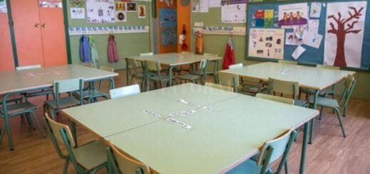 alt="un aula vacía, con cuatro mesas y varias sillas cada una, sobre una pared están pegados dibujos de niños"