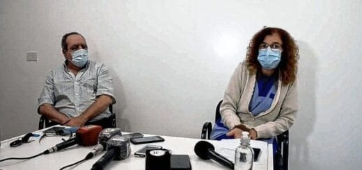 alt="en primer plano, micrófonos de radio y televisión sobre una mesa, detrás de la mesa, sentados, un hombre y una mujer"