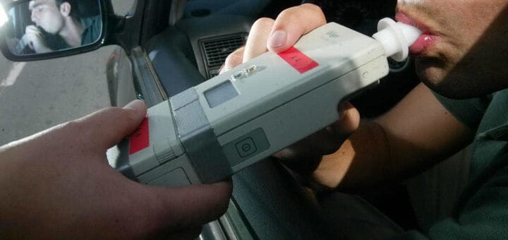 alt="en la fotografía, una mano sostiene un alcoholímetro que está siendo soplado por una persona que va en un auto"