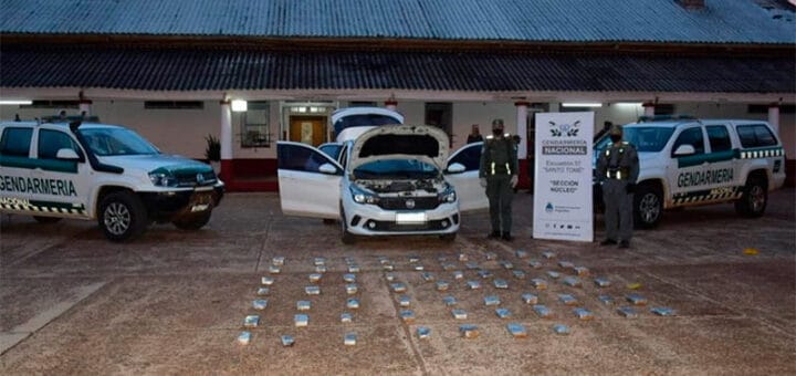 alt="en primer plano, en el suelo, varios envoltorios con marihuana: detrás, tres vehículos, el del centro es un auto secuestrado donde transportaban la droga, a sus lados dos camionetas de la Gendarmería Nacional: detrás de todo, un inmueble"