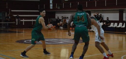 alt="en la imagen, dos jugadores con vestimenta verde, uno de los cuales tiene en sus manos un balón, junto a ellos un jugador con vestimenta blanca; todos están en un estadio de basquet, con tribunas casi vacías"