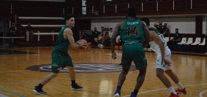 alt="en la imagen, dos jugadores con vestimenta verde, uno de los cuales tiene en sus manos un balón, junto a ellos un jugador con vestimenta blanca; todos están en un estadio de basquet, con tribunas casi vacías"