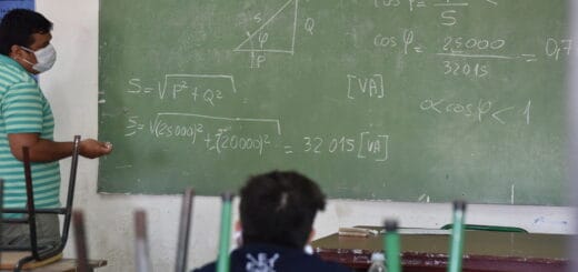 alt="un profesor, con barbijo, frente a un pizarrón sobre el que hay ejercicios matemáticos, mientras un alumno observa"