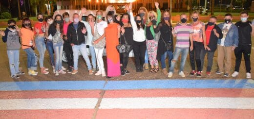 alt="en primer plano, una senda peatona pintada con los colores alusivos a la comunidad trans; al fondo, un grupo de personas que pintó la senda peatonal"