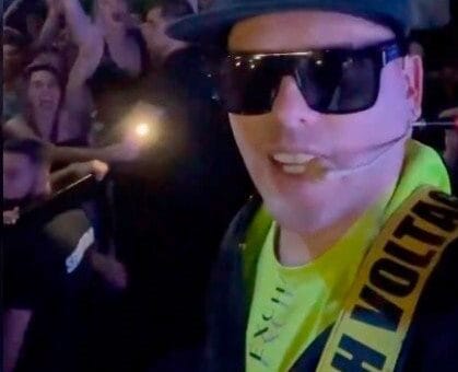 alt="un cantante, con gorrita, lentes negros, una cinta que le cruza el hombro; detrás suyo, público saltando y bailando, muy cerca unas personas de otras"