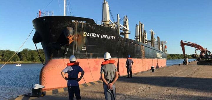 alt="un barco amarrado a un muelle, en un río. Dos hombres observan la nave"