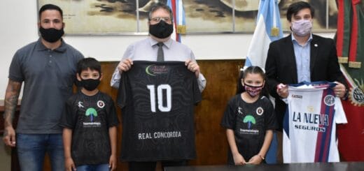 alt="en la foto, tras hombres y dos niños, dos de los mayores sostienen camisetas de fútbol, detrás de ellos un cuadro que ocupa gran parte de la pared"