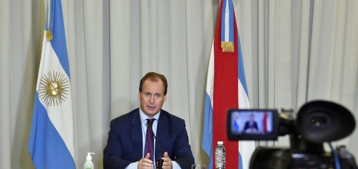 alt="en la imagen, habla el gobernador de entre ríos ante una cámara de televisión, al lado del gobernador las banderas argentina y entrerriana, detrás una gran cortina blanca"