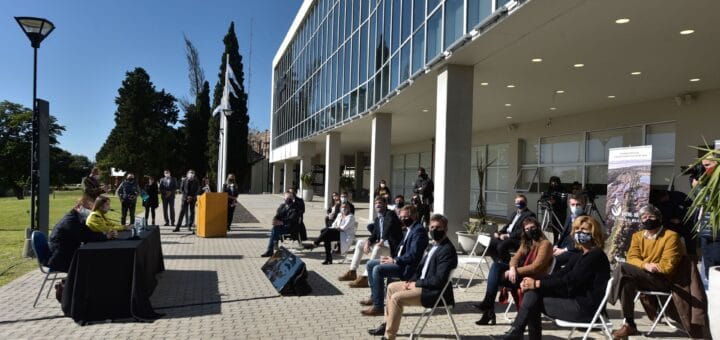 alt="en la fotografía, a la derecha, varias persoanas sentadas en sillas al aire libre, debajo de la entrada de un edificiio; a la izquierda, en una mesa principal, sentadas dos personas"