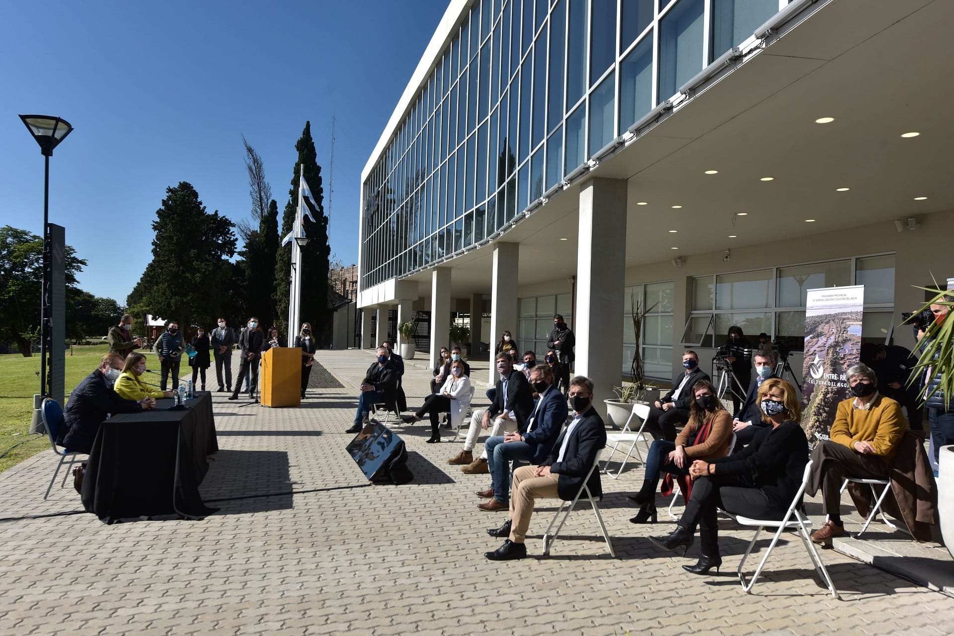 alt="en la fotografía, a la derecha, varias persoanas sentadas en sillas al aire libre, debajo de la entrada de un edificiio; a la izquierda, en una mesa principal, sentadas dos personas"