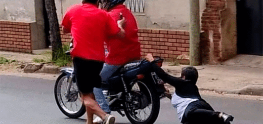 alt="una mujer se toma de la parte de atrás de una moto, el motociclista está en marcha, otra persona intenta subirse al rodado a la carrera"