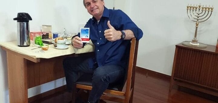 alt="en la foto puede verse a un hombre sentado en un escritorio, exhibiendo un sobre, sobre el escritorio hay varios elementos; a la derecha, sobre un mueble hay un candelabro de nueve brazos"