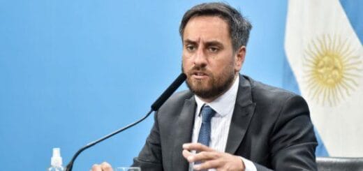 alt="en la fotografía puede verse a un hombre sentado, frente a un micrófono, en el fondo una bandera argentina"