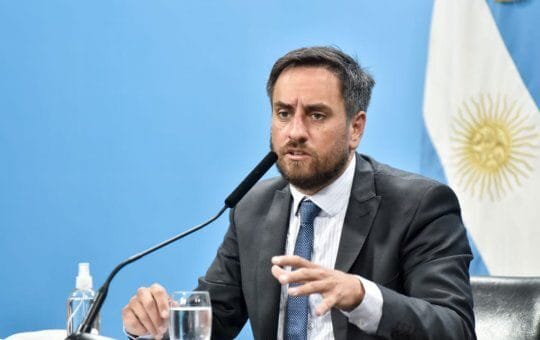 alt="en la fotografía puede verse a un hombre sentado, frente a un micrófono, en el fondo una bandera argentina"