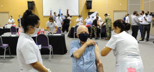 alt="en la foto puede verse, en primer plano, a una enfermera aplicando una vacuna a un hombre mientras otra enfermera observa, al fondo varias personas en un salón"