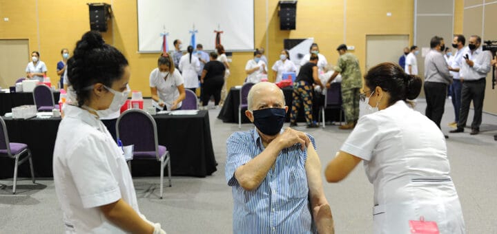 alt="en la foto puede verse, en primer plano, a una enfermera aplicando una vacuna a un hombre mientras otra enfermera observa, al fondo varias personas en un salón"