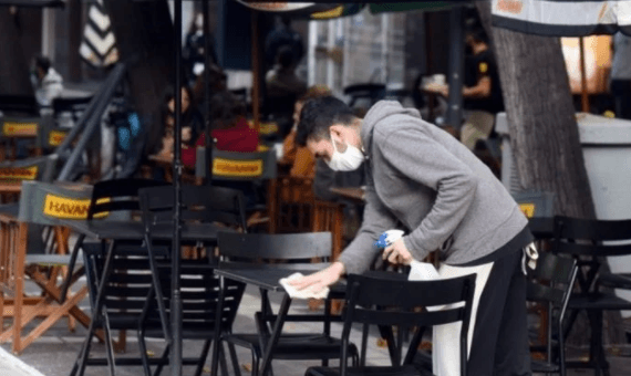 alt="en la imagen puede verse a una persona con barbijo limpiando una mesa en un restaurante vacío"
