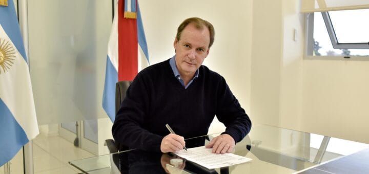alt="en la imagen, el gobernador gustavo bordet firma unos papeles, detrás de él una bander de Entre Ríos"