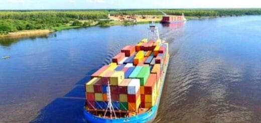 alt="en primer plano puede verse un barco con contenedores navegando por un río, al fondo un puerto con un barco amarrado"