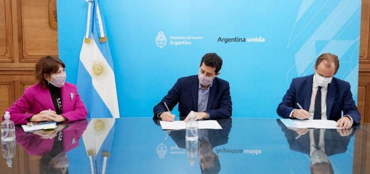 alt="dos hombres firman papeles sobre una mesa, una mujer los observa, detrás de ellos una bandera argentina"