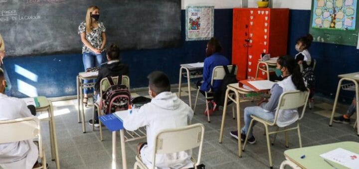 alt="en la foto pueden verse en primer plano varios niños sentados en bancos en una escuela, al fondo una docente y detrás de ella un pizarrón"