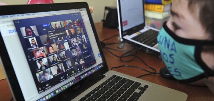 alt="en la imagen puede verse, en primer plano, un niño frente a una pantalla de computadora, más atrás se ve otra pantalla de computadora"