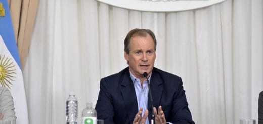 alt="en la imagen, un hombre habla a un micrófono, a su izquierda, un vaso, una botella de agua y una botella de alcohol"