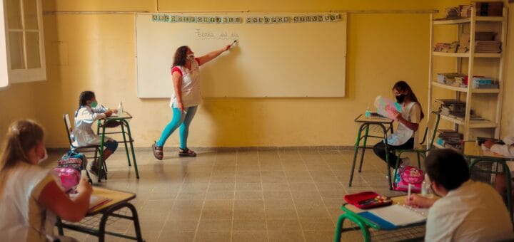 alt="en la imagen, una maestra da clases frente a sus alumnos"