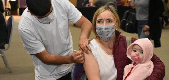 alt="en la imagen puede verse a un enfermero vacunando a una mujer que tiene un bebé en los brazos"