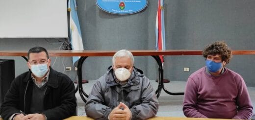 alt="en la imagen se ve a tres personas sentadas ante una mesa en la que hay sobres, detrás de las personas hay un cartel con la inscripción cafesg y a sus lados dos banderas"