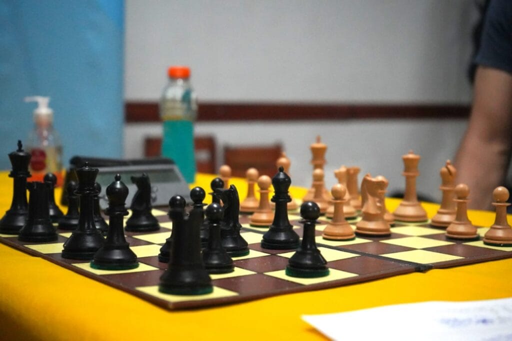 alt="sobre una mesa puede verse un tablero de ajedrez, las piezas del juego, un cronómetro, una botella con un líquido azul y un frasco de alcohol en gel, a un costado se ve el brazo de una persona"