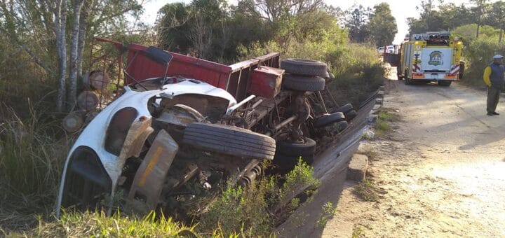alt="en la imagen puede verse a un camión, cargado con troncos, volcado sobre uno de sus laterales tras caer de un puente; más atrás hay un persona y después un vehículo del Cuerpo de Bomberos Voluntarios de Concordia"