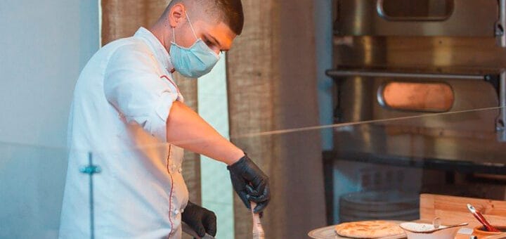 alt="en la imagen puede verse a una persona con barbijo preparando una pizza en un local gastronómico"