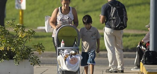 alt="en la imagen, una mujer empuja un coche de bebés mientras es acompañada por un niño, a su derecha y detrás de de ella un hombre con una mochila está parado, hay otro que está sentado al lado de una columna"