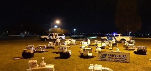 alt="en la imagen puede verse varios bultos con mercaderías incautadas, al fondo se ven dos móviles de la Prefectura Naval Argentina y un uniformado que toma notas"